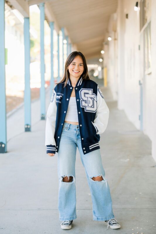 high school senior in a varsity jacket and ripped jeans smiles in a school hallway, perfect for high school senior portraits.