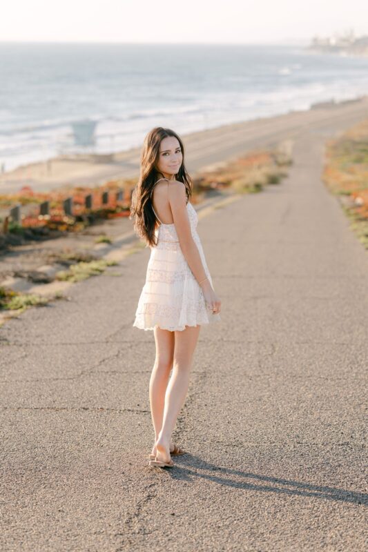 Woman in a white dress stands on a coastal path, perfect for senior portraits San Diego, ocean waves behind her.