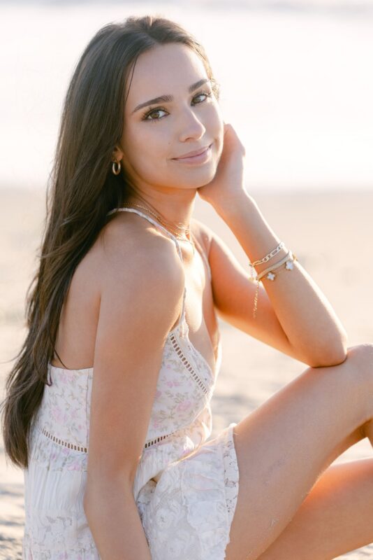 Woman with long brown hair in a floral dress on the beach, smiling—perfect for senior portraits San Diego style.