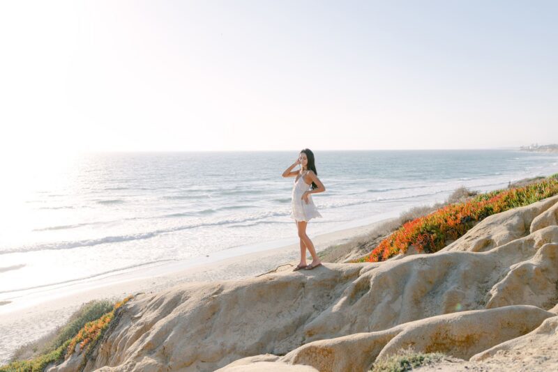 Woman in a white dress stands on a rocky cliff overlooking the ocean—perfect for senior portraits San Diego style.