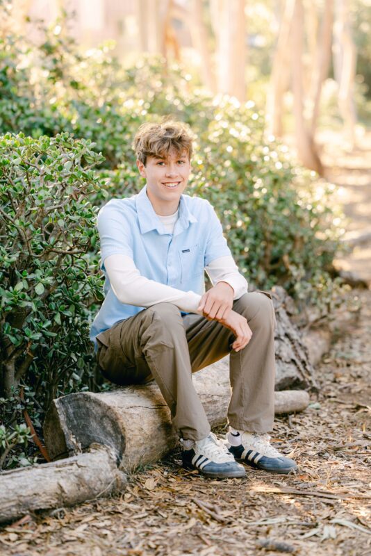 Smiling young man sits on a fallen log outdoors—perfect inspiration for senior photos for guys San Diego.