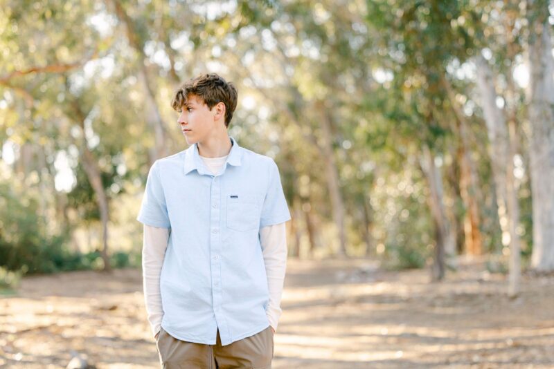 Young man in a light blue shirt stands in a sunlit forest—perfect for senior photos for guys San Diego.