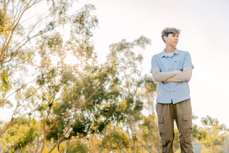 Young person with arms crossed stands outdoors among trees—perfect for senior photos for guys in San Diego.