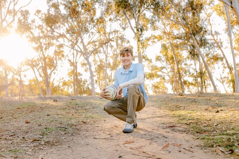 Smiling young person kneels on a dirt path with a basketball—perfect for senior photos for guys San Diego.