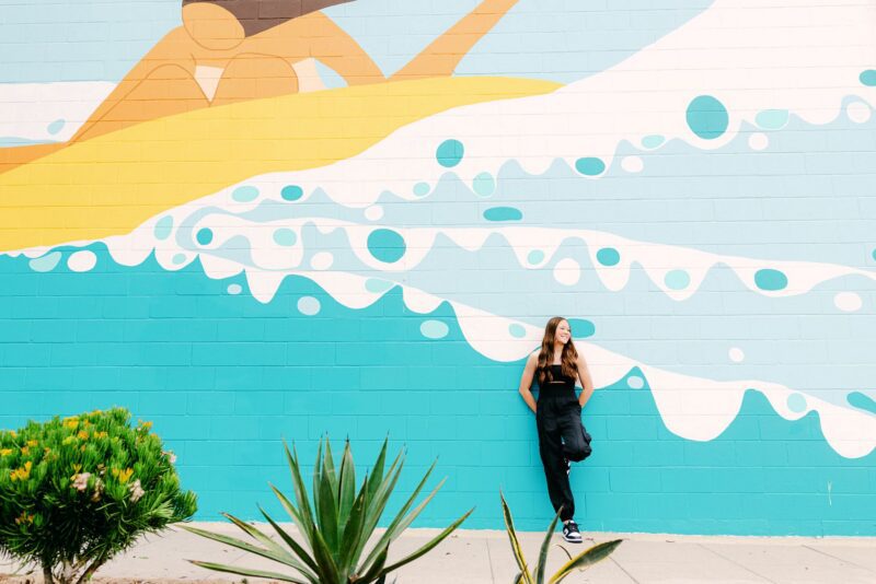 Senior Portraits Ideas: High school senior girl stands by a vibrant mural of a surfer on waves, plants in the foreground, sun shining.