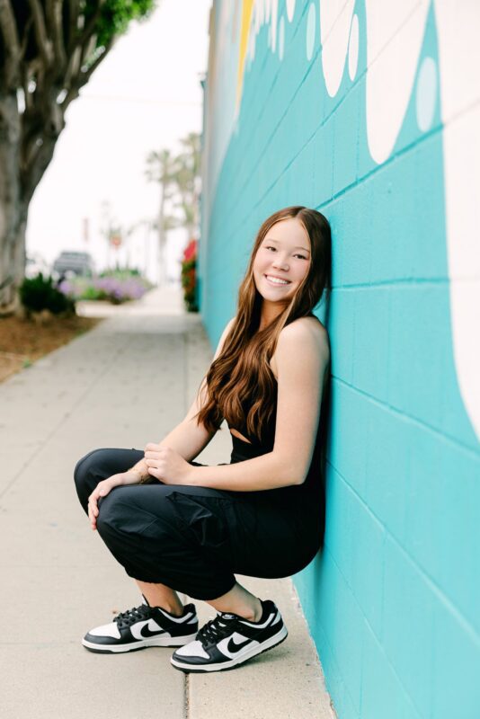 High school senior girl with long hair squats and smiles by a blue mural wall—great inspiration for Senior Portraits Ideas.