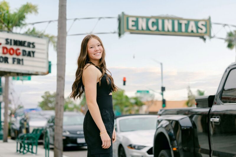 Smiling High school senior girl stands on a street in Encinitas—perfect for creative Senior Portraits Ideas with the iconic sign in view.