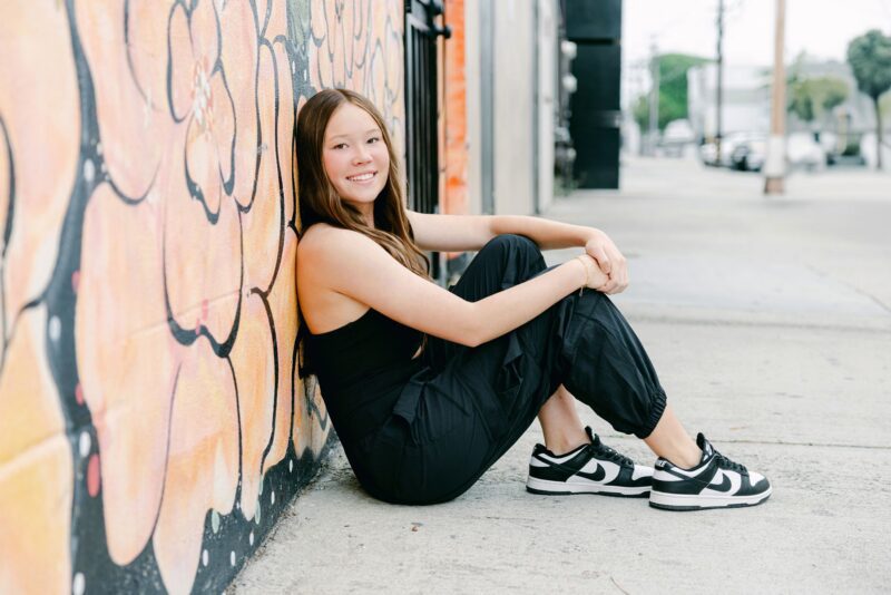 High school senior girl in black outfit smiles against a colorful mural—unique Senior Portraits Ideas for vibrant city settings.