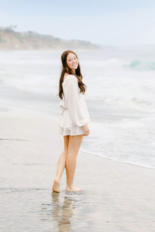 High school, senior girl in a white dress stands barefoot on the beach—capturing unique Senior Portraits Ideas by the shoreline.