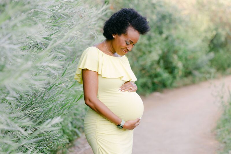 A pregnant woman in a yellow dress smiles, holding her belly on a nature path by Carlsbad Maternity Photographer.
