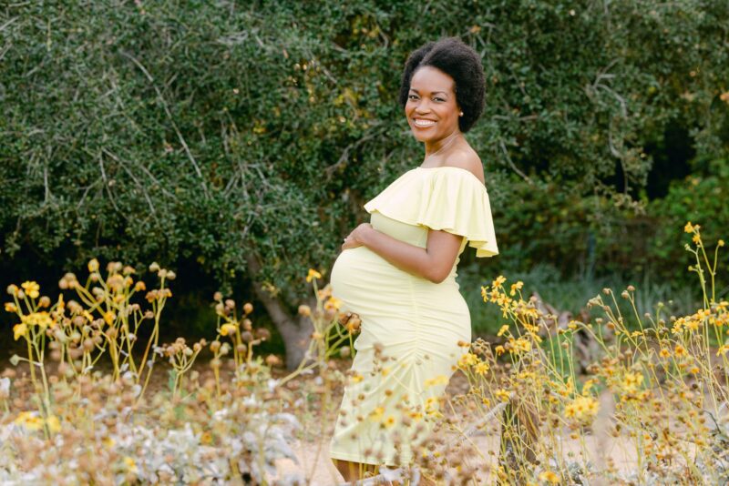 Smiling pregnant woman in yellow dress stands in wildflower field—captured by Carlsbad Maternity Photographer.