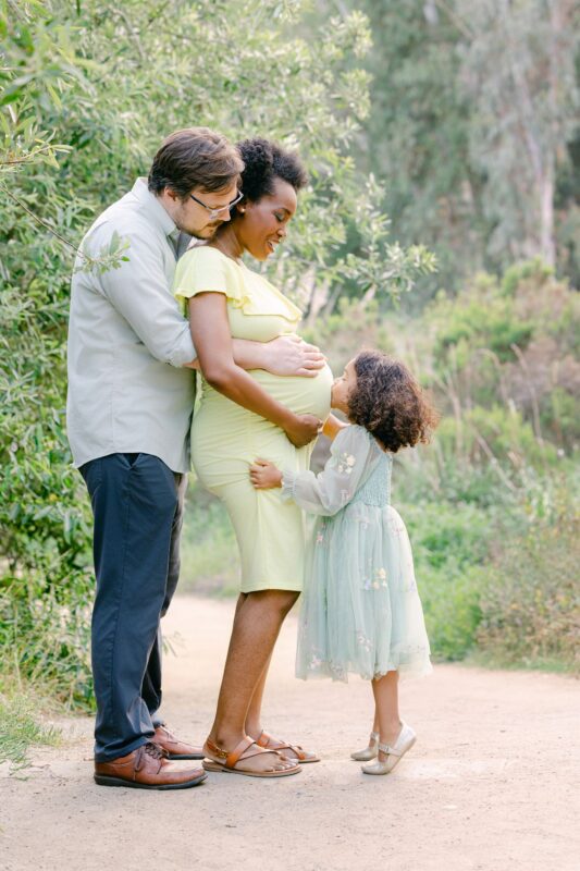 A couple stands on a path as their young daughter kisses mom's pregnant belly—Carlsbad Maternity Photographer.