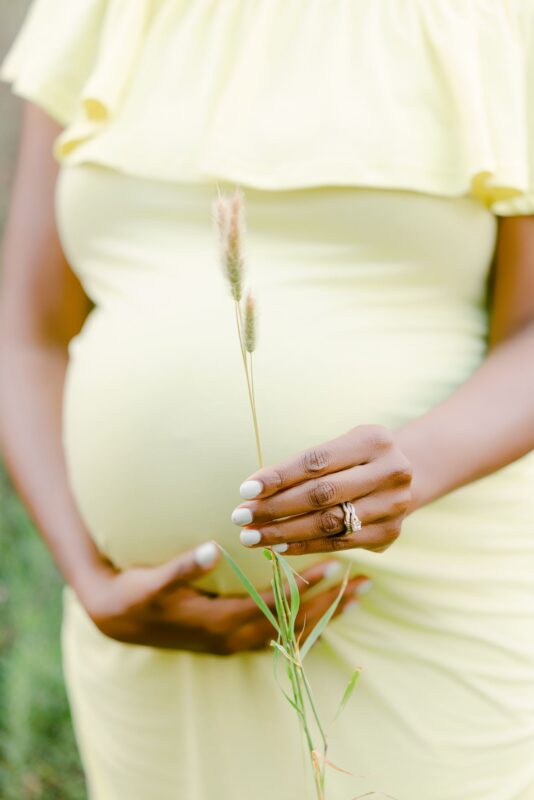A pregnant person in yellow cradles their belly and grass sprig—captured by Carlsbad Maternity Photographer.