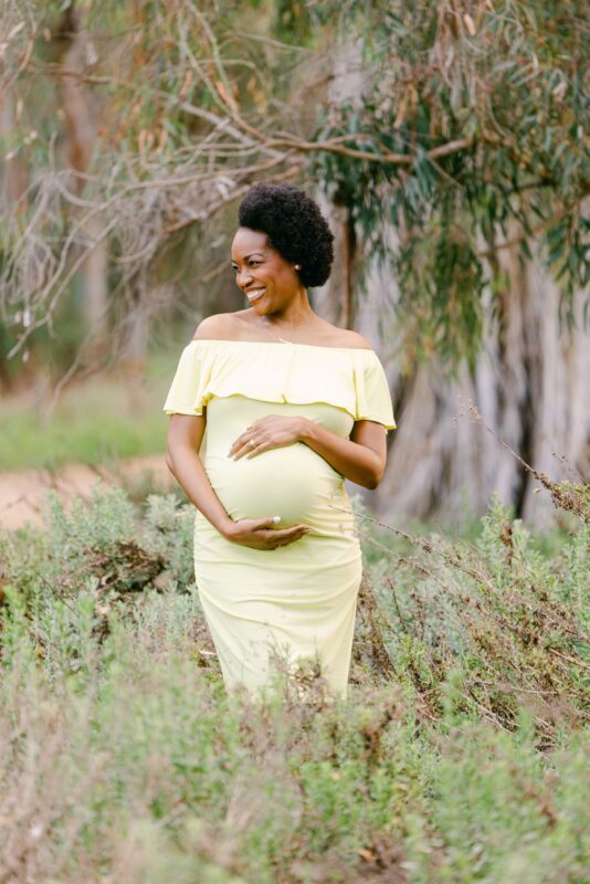 Smiling pregnant woman in yellow dress stands outdoors, holding her belly—captured by a Carlsbad Maternity Photographer.