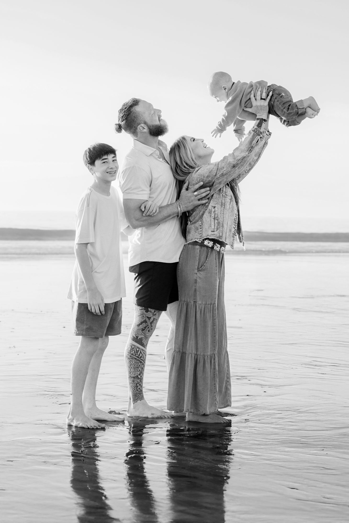 Father lifting his baby in the air while the family laughs on the beach, black and white
