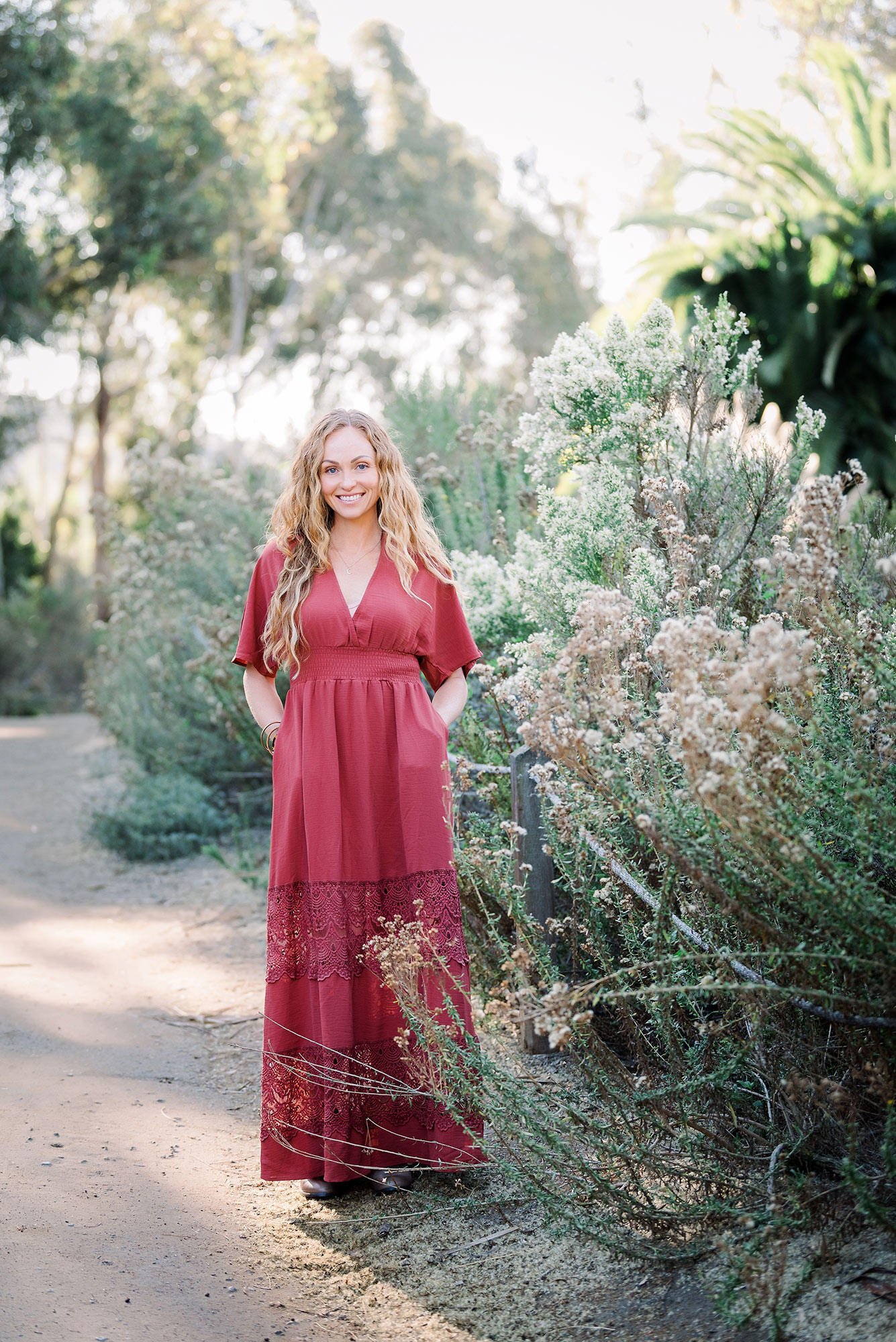 A woman in a long red dress stands on a garden path, surrounded by green and flowering bushes.