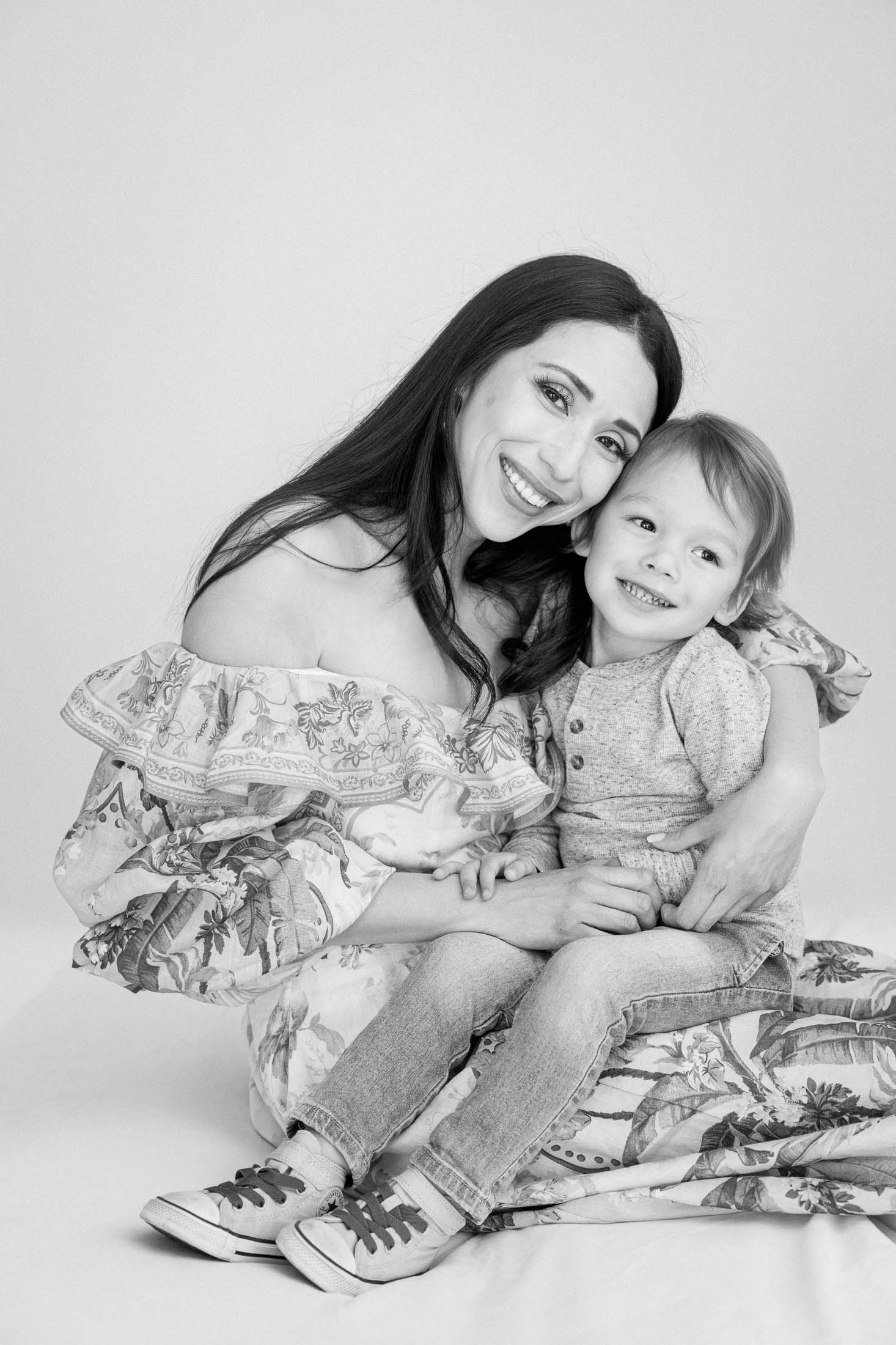 A woman smiling and hugging a young child, both sitting together and looking at the camera in a joyful moment.