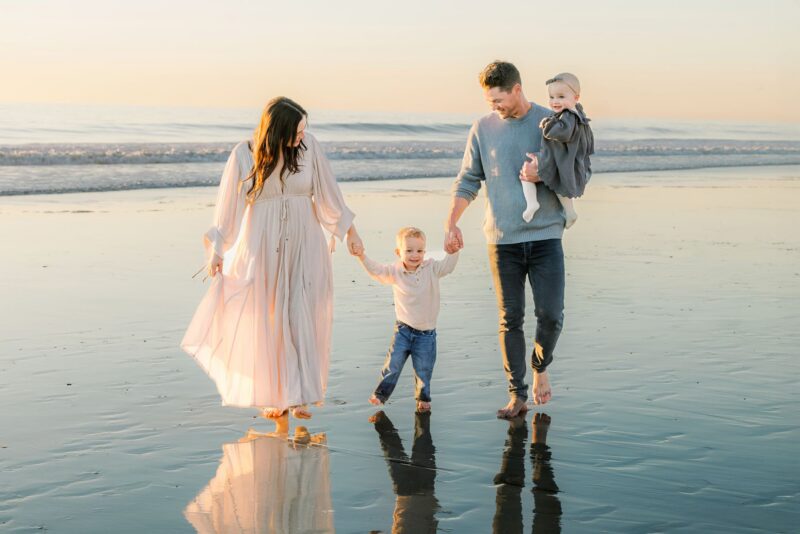A San Diego Family Photographer captures a family of four walking barefoot on the beach at sunset, holding hands and smiling.