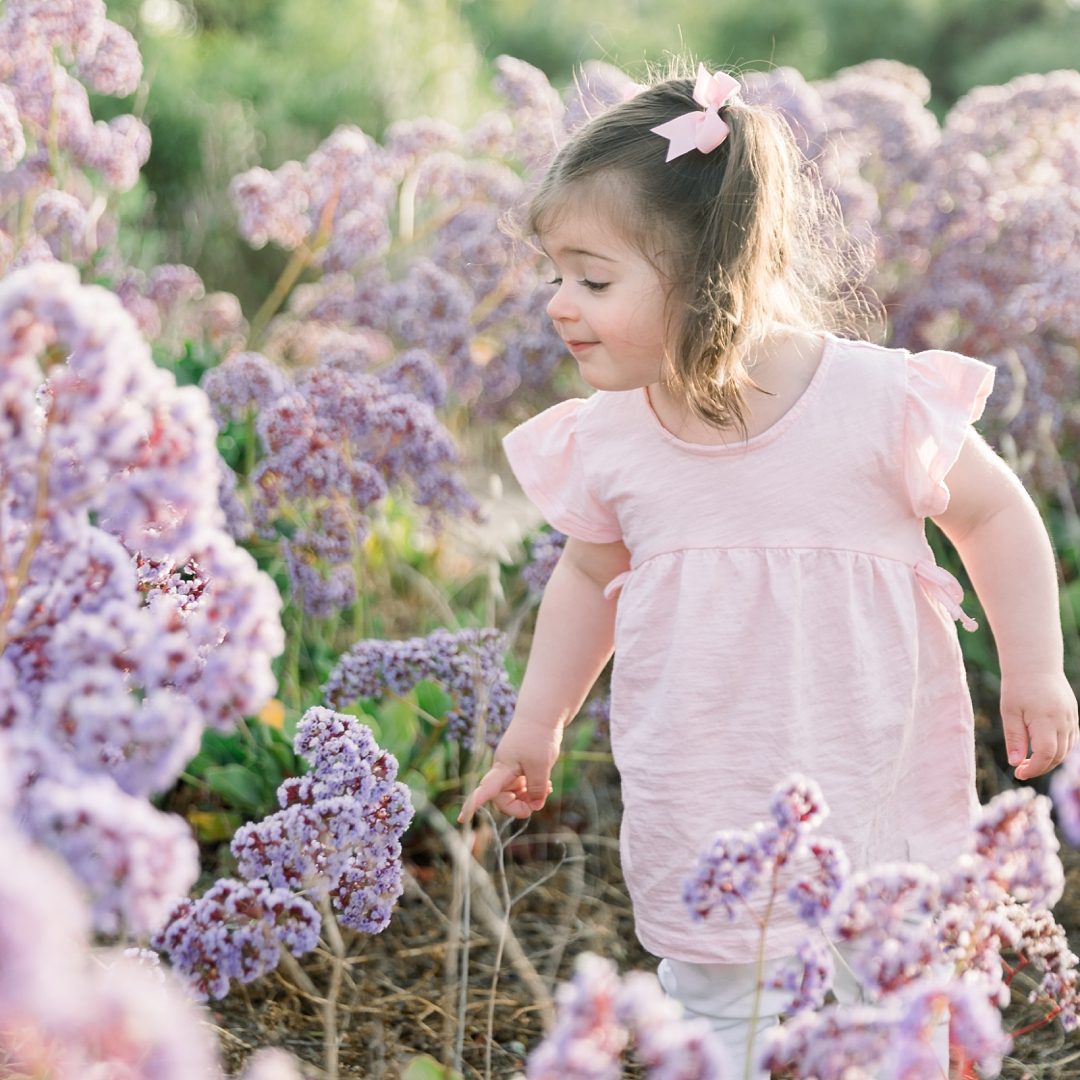 Little girl in a pink dress explores a garden filled with blooming purple flowers on a sunny day.