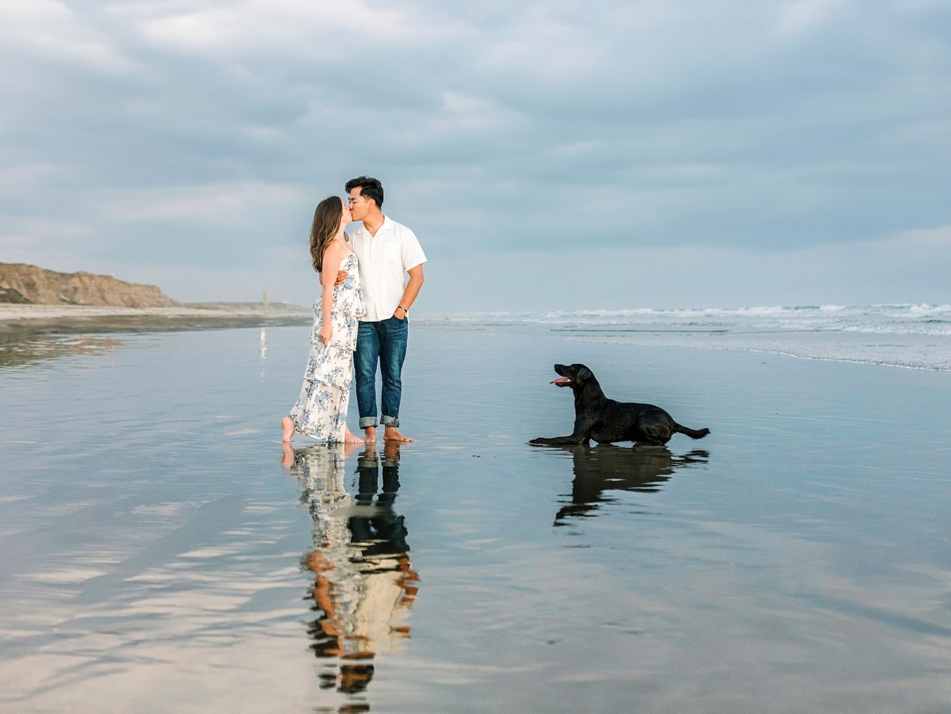 A couple stands barefoot on the beach, facing each other, with a black dog lying nearby on the wet sand.