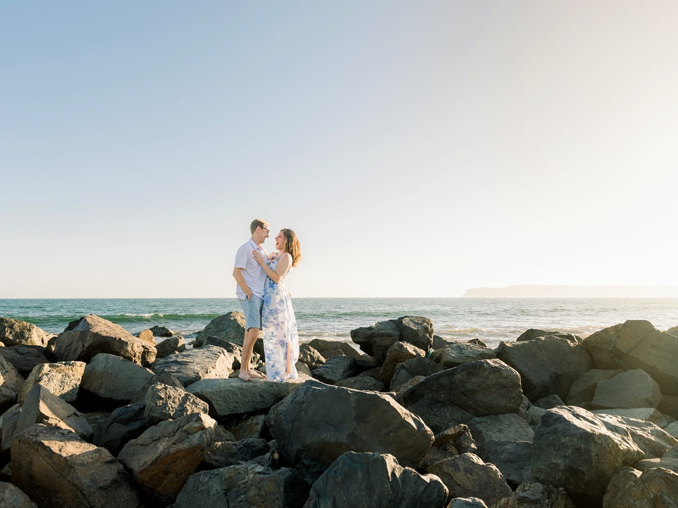 A couple stands on large rocks by the ocean, smiling at each other under a clear sky with the sun shining.