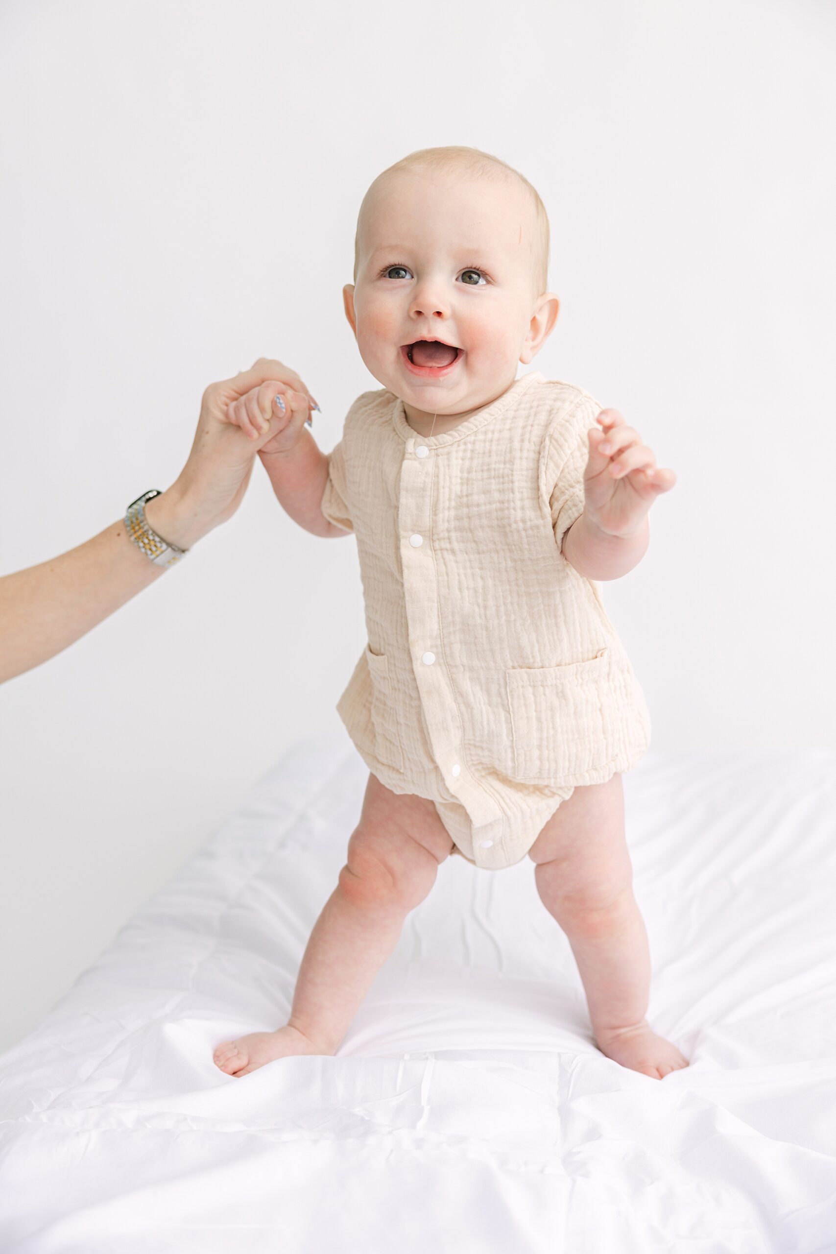 A baby in a tan romper stands on a white surface, holding an adults hand for balance.