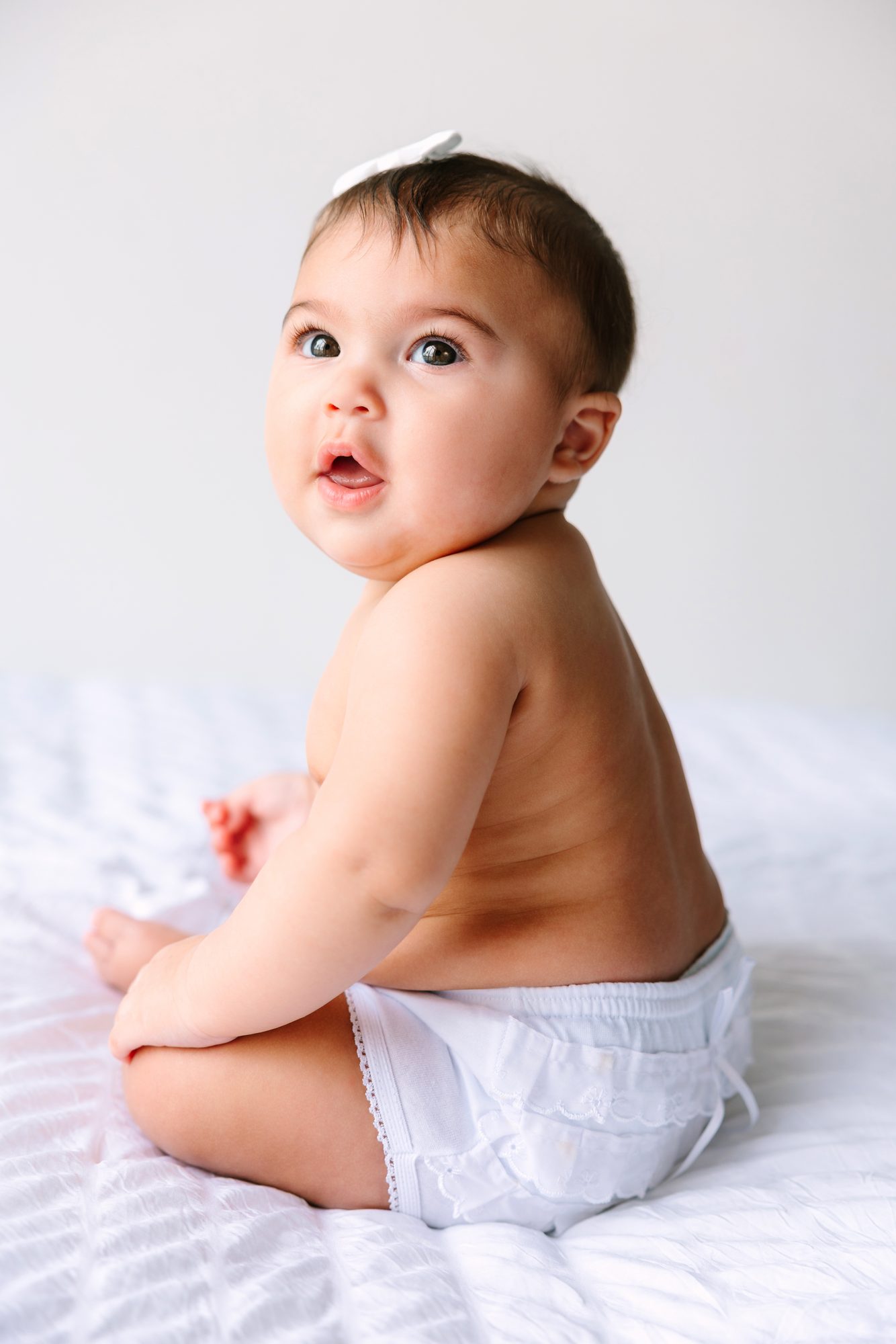 Baby girl sitting on a white bed, looking back over her shoulder