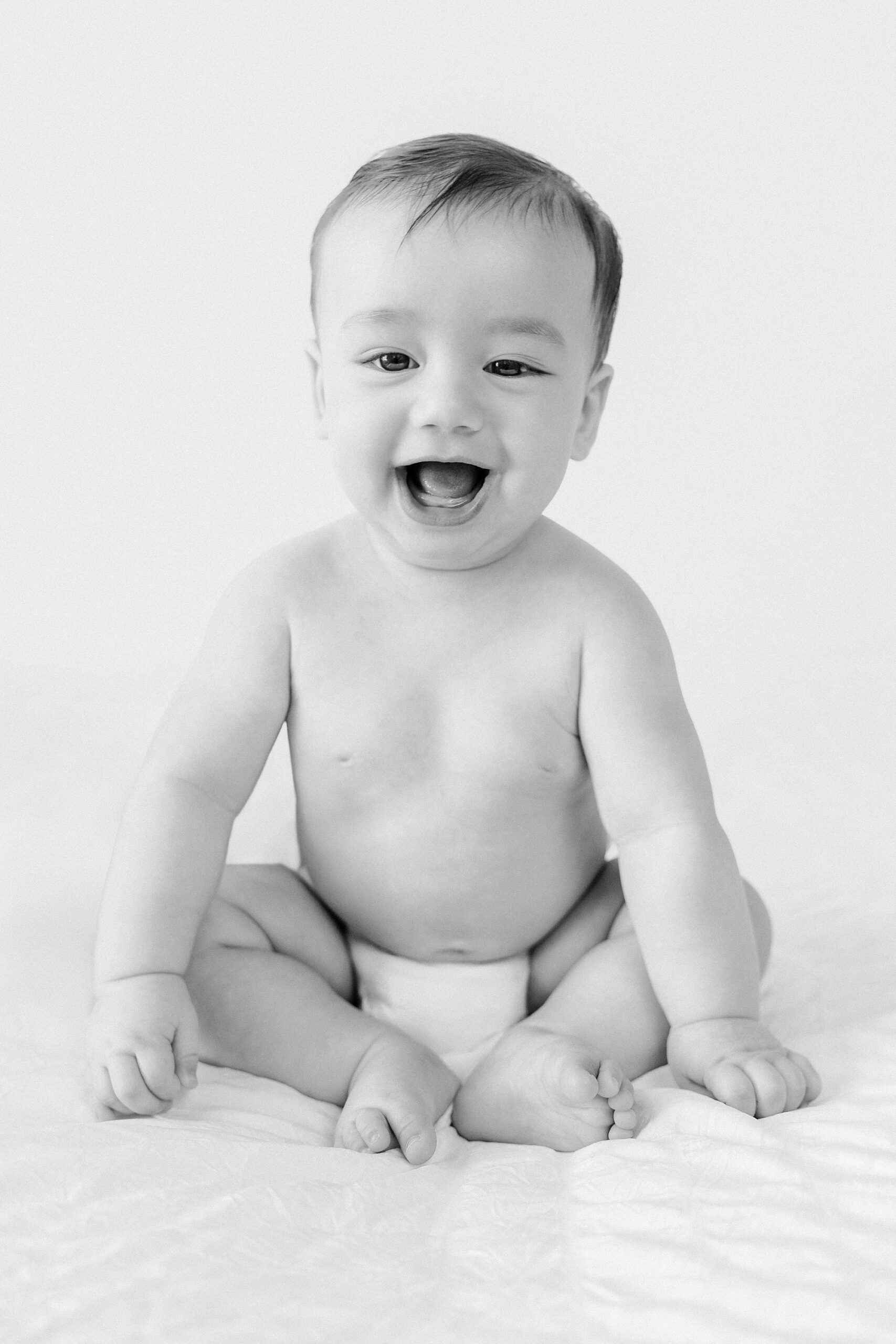 A laughing baby in a diaper sitting on a soft surface.