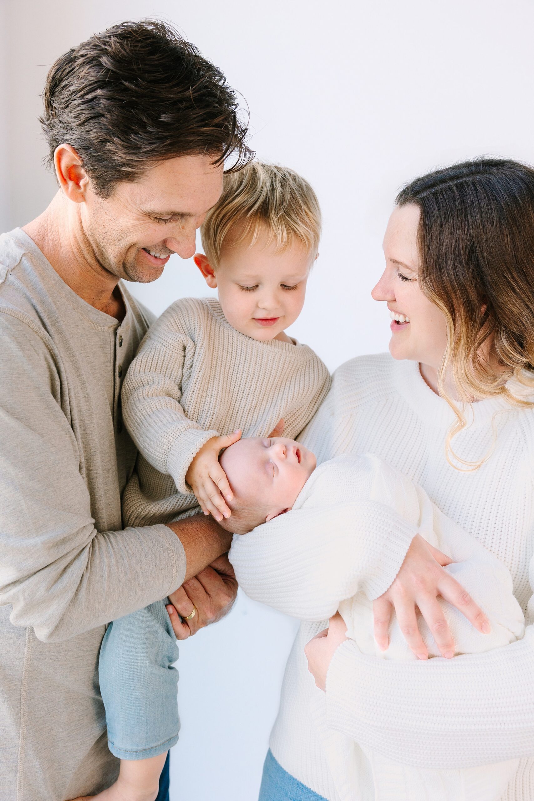 A man and woman smiling at a sleeping newborn held in the womans arms, while a toddler touches the babys head.