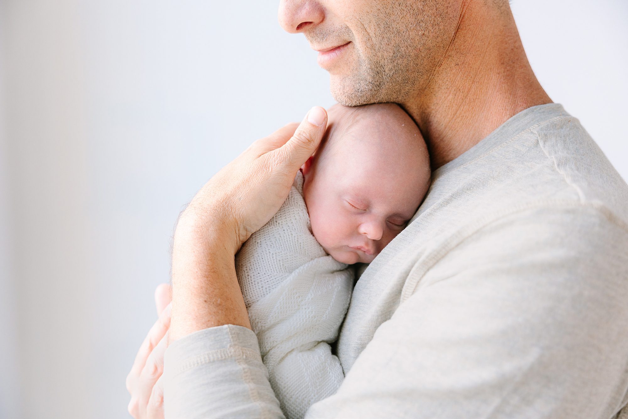 A man cradles a swaddled newborn baby sleeping against his chest.