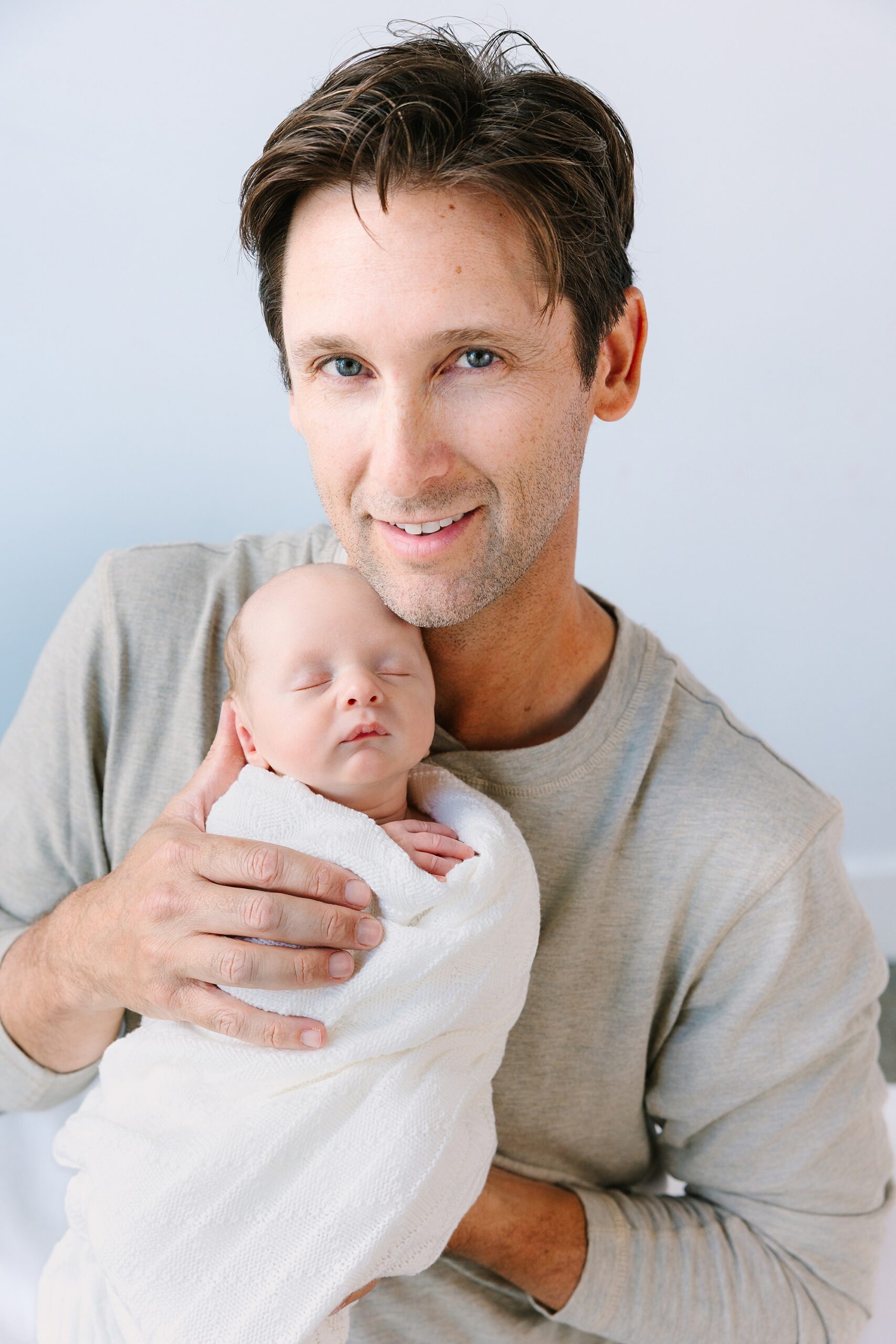 A man holding a newborn baby wrapped in a white blanket against his cheek.
