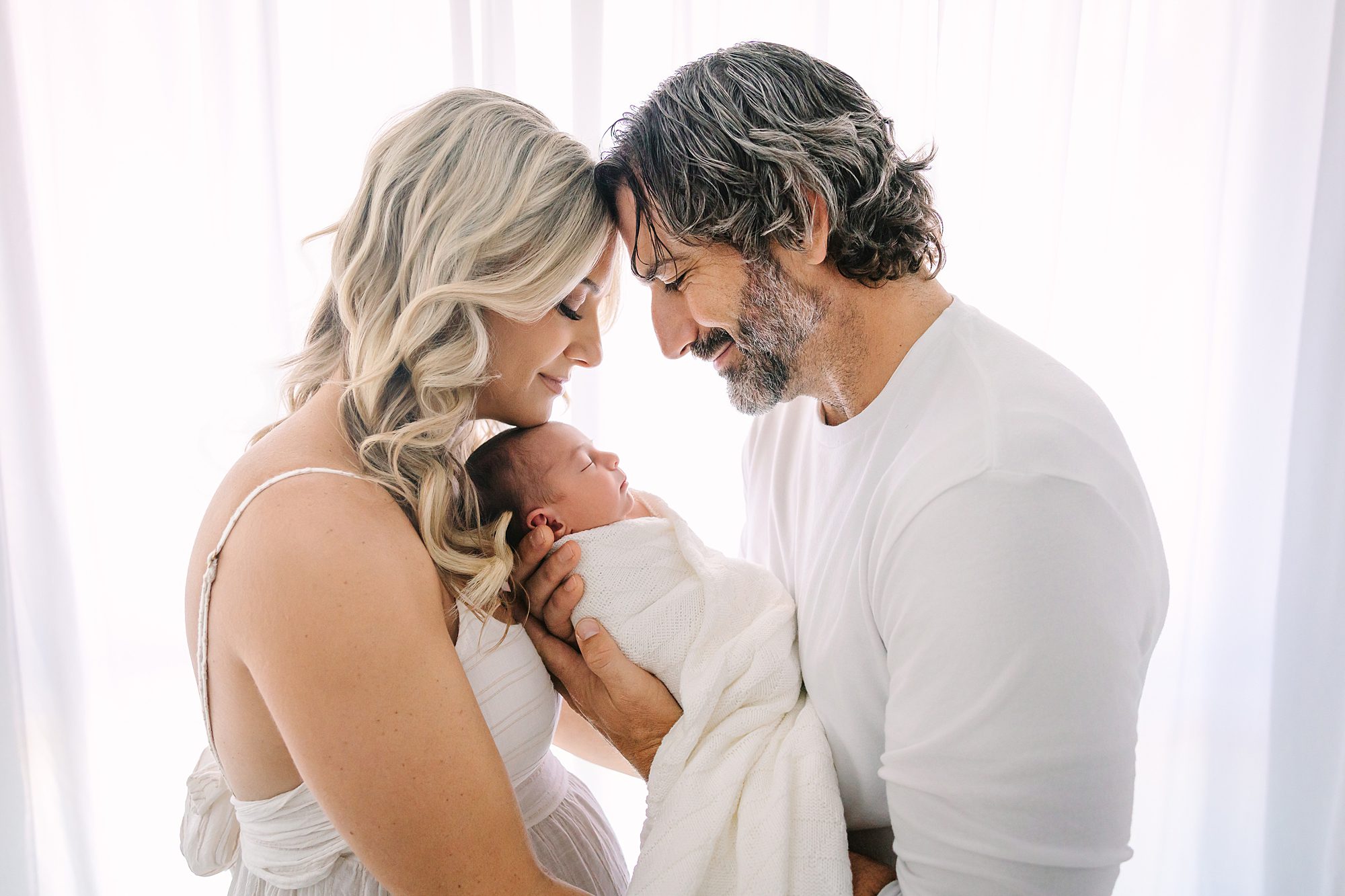 A mother and father hold their newborn baby, all touching foreheads.