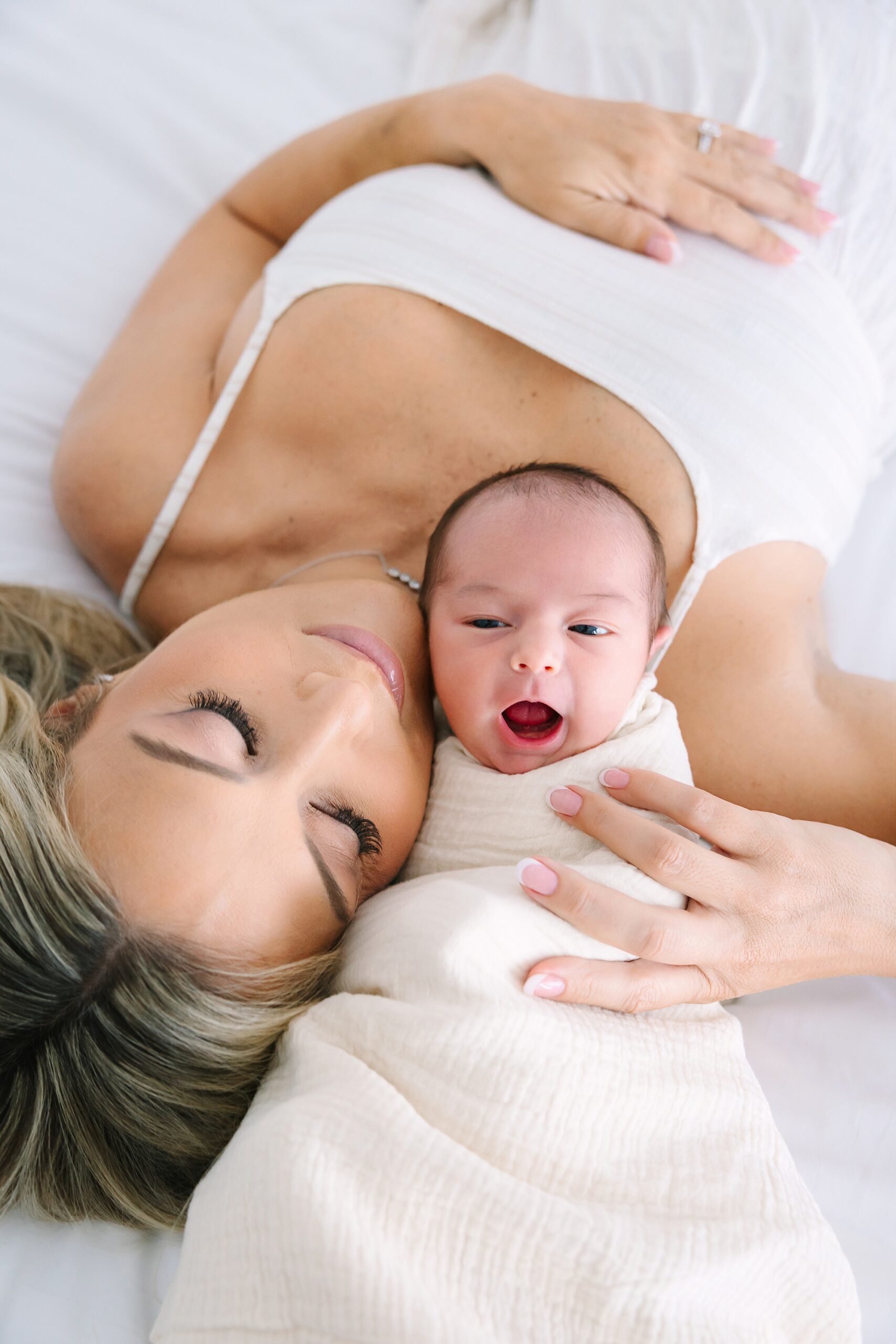 A mother lying on a bed rests her cheek against her swaddled newborn baby.