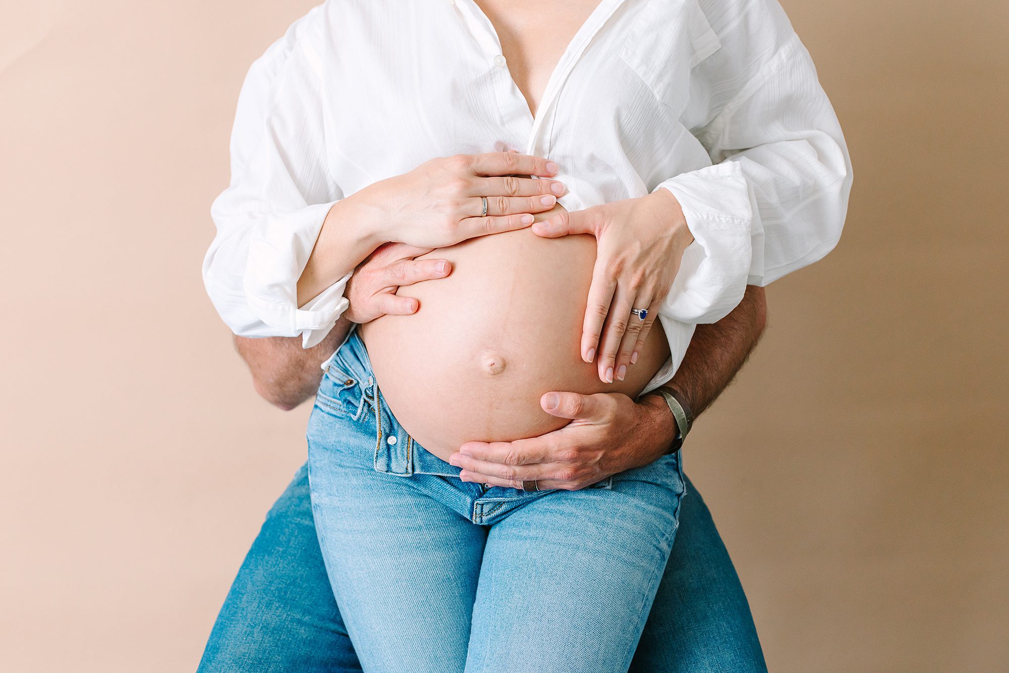 A pregnant person in jeans and a white shirt with hands cradling their belly, supported by a partners arms.