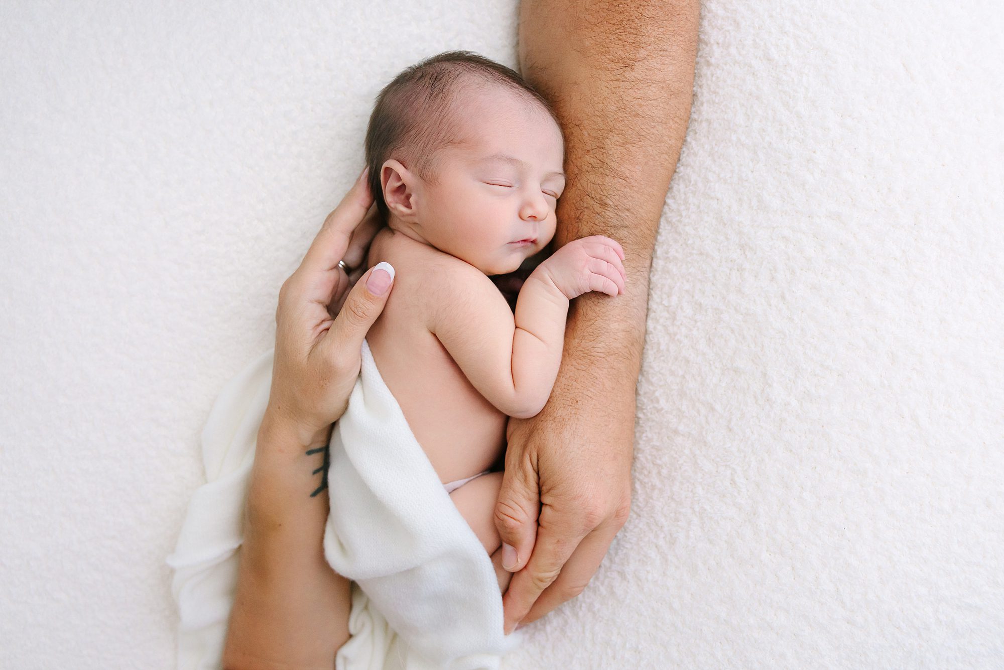 A sleeping newborn baby held in the hands of two adults.