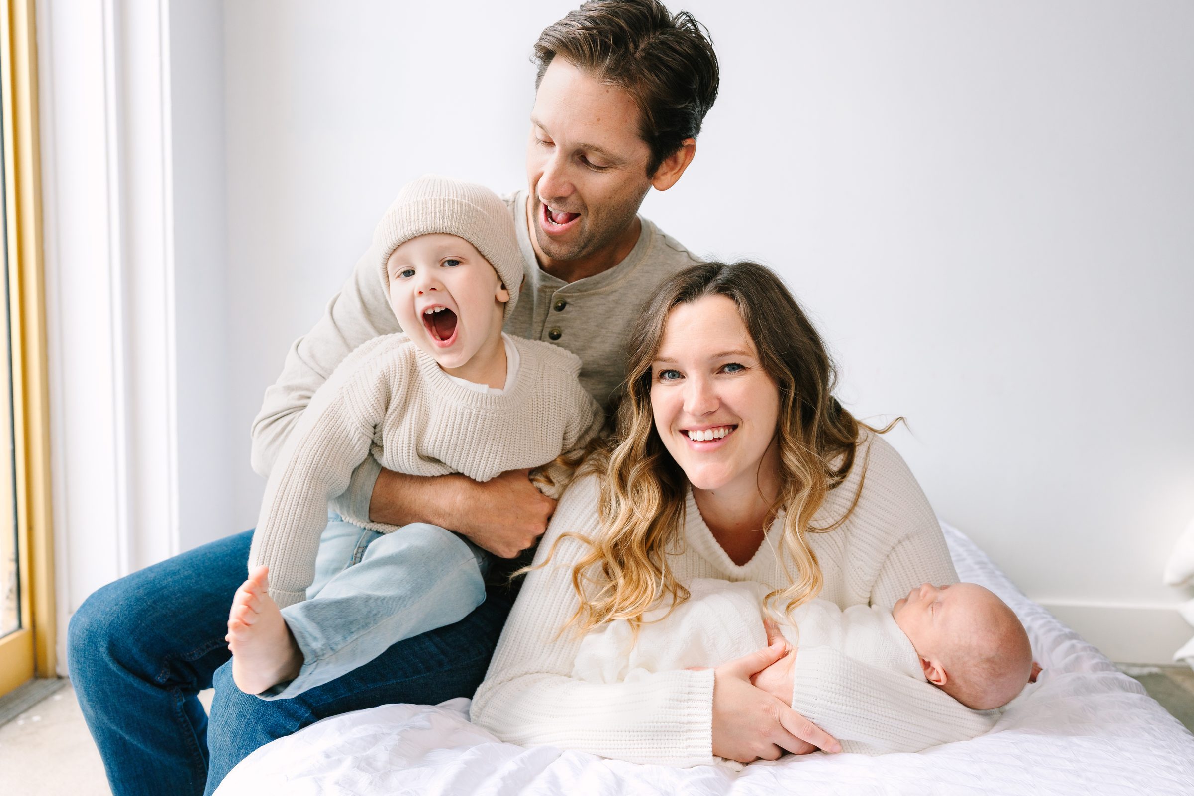 Family of four laughing together on a white bed in the Vallentyne Photography studio in Carlsbad
