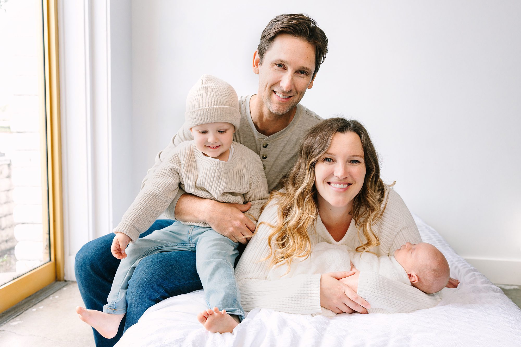 A smiling family of four sitting together on a white bed.