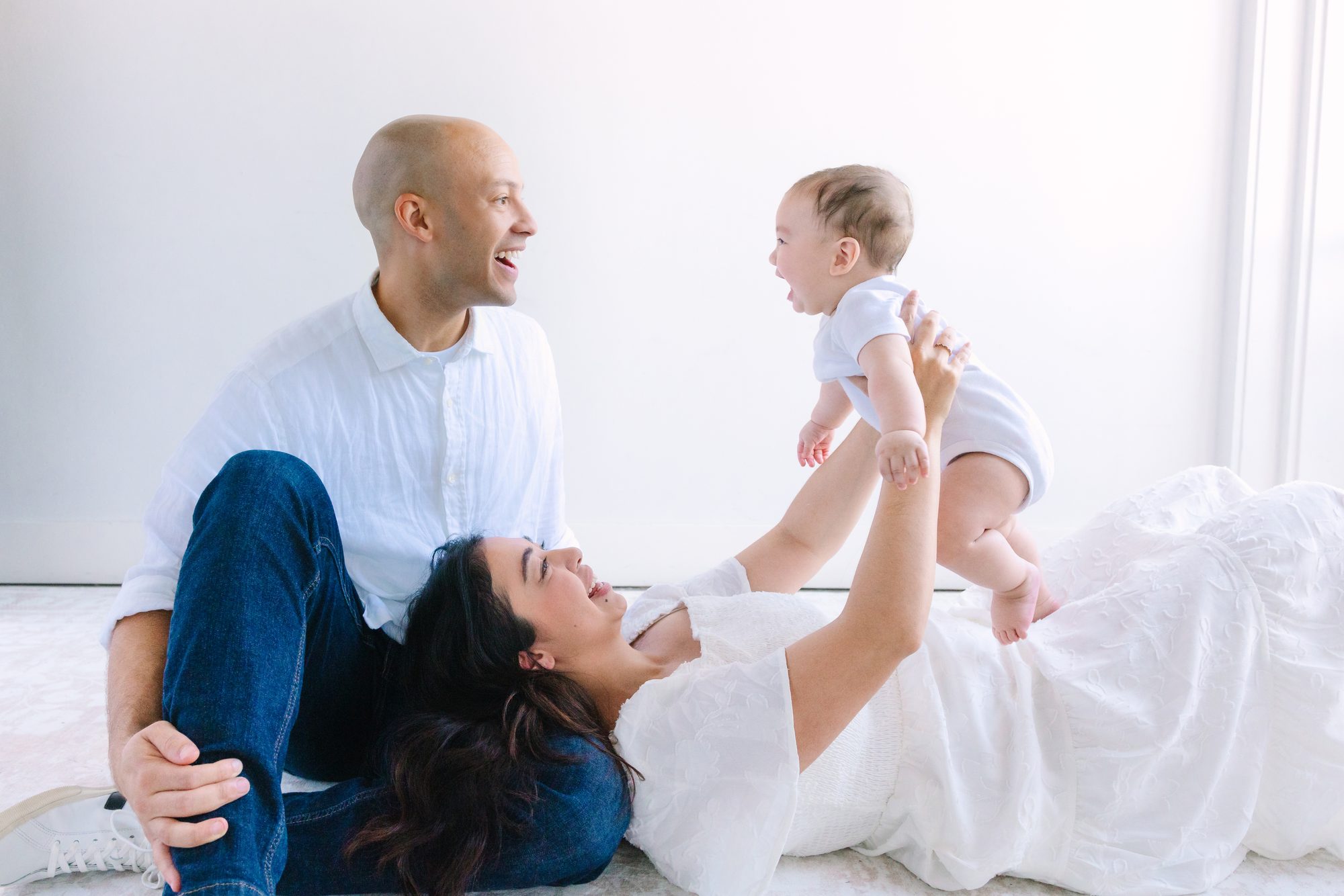 Mother lying on the floor lifting her baby while dad laughs, playful studio family photo