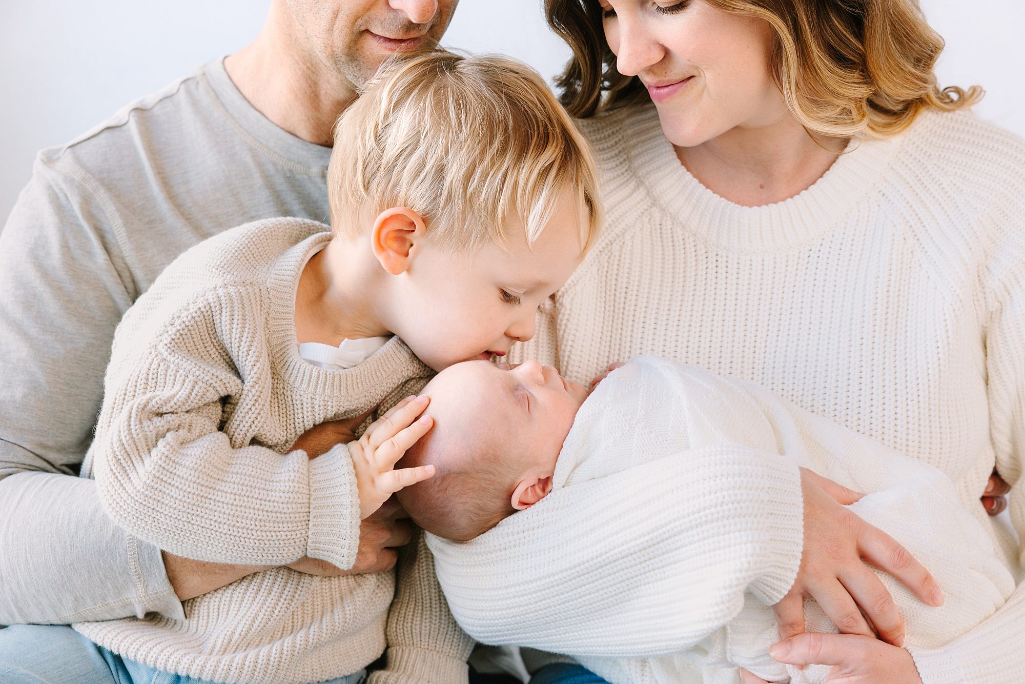 A young boy kisses the forehead of a newborn baby held by their parents.