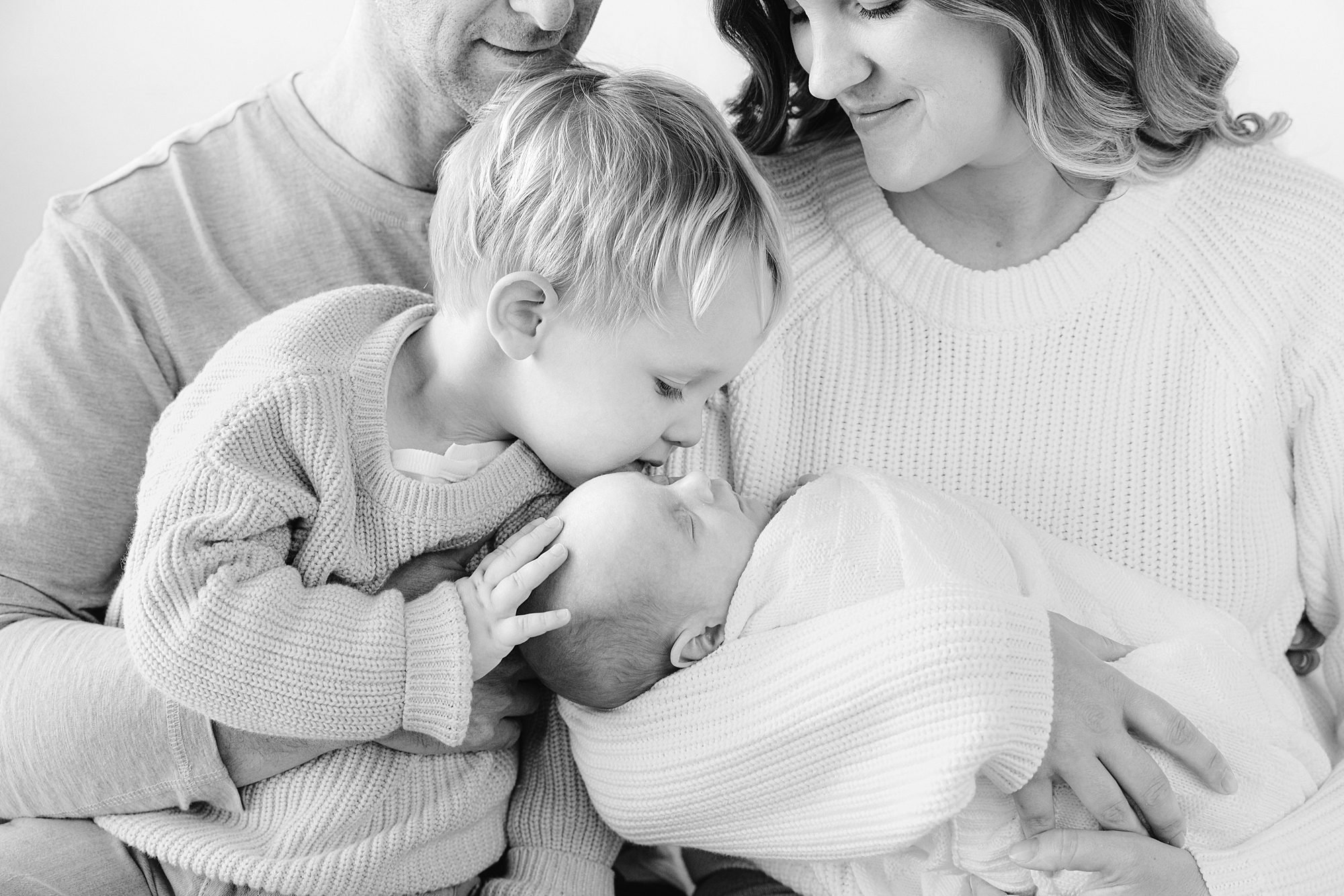 A young child kisses a newborn baby held by their parents.