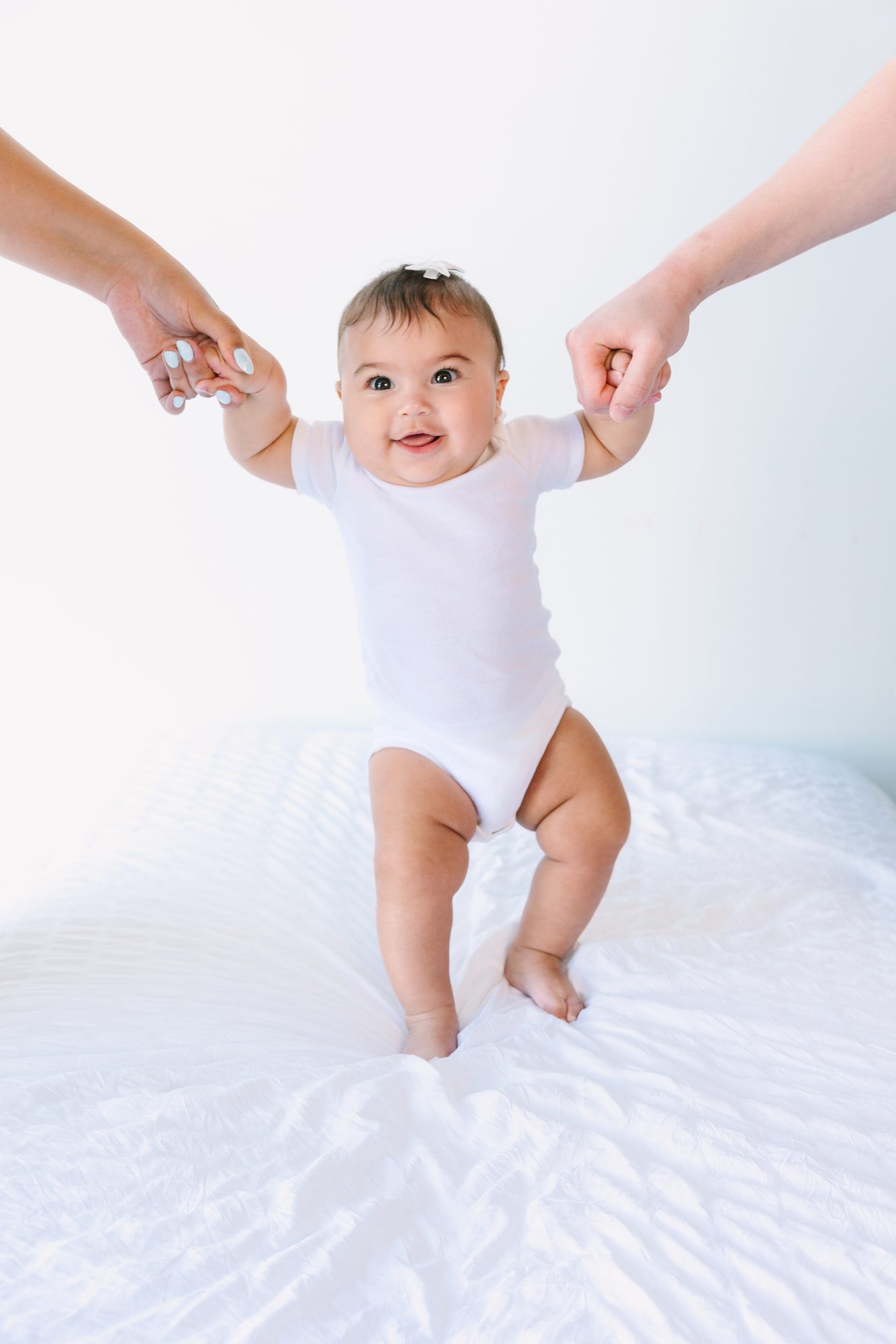 Baby girl taking steps on a white bed while holding her parents' hands