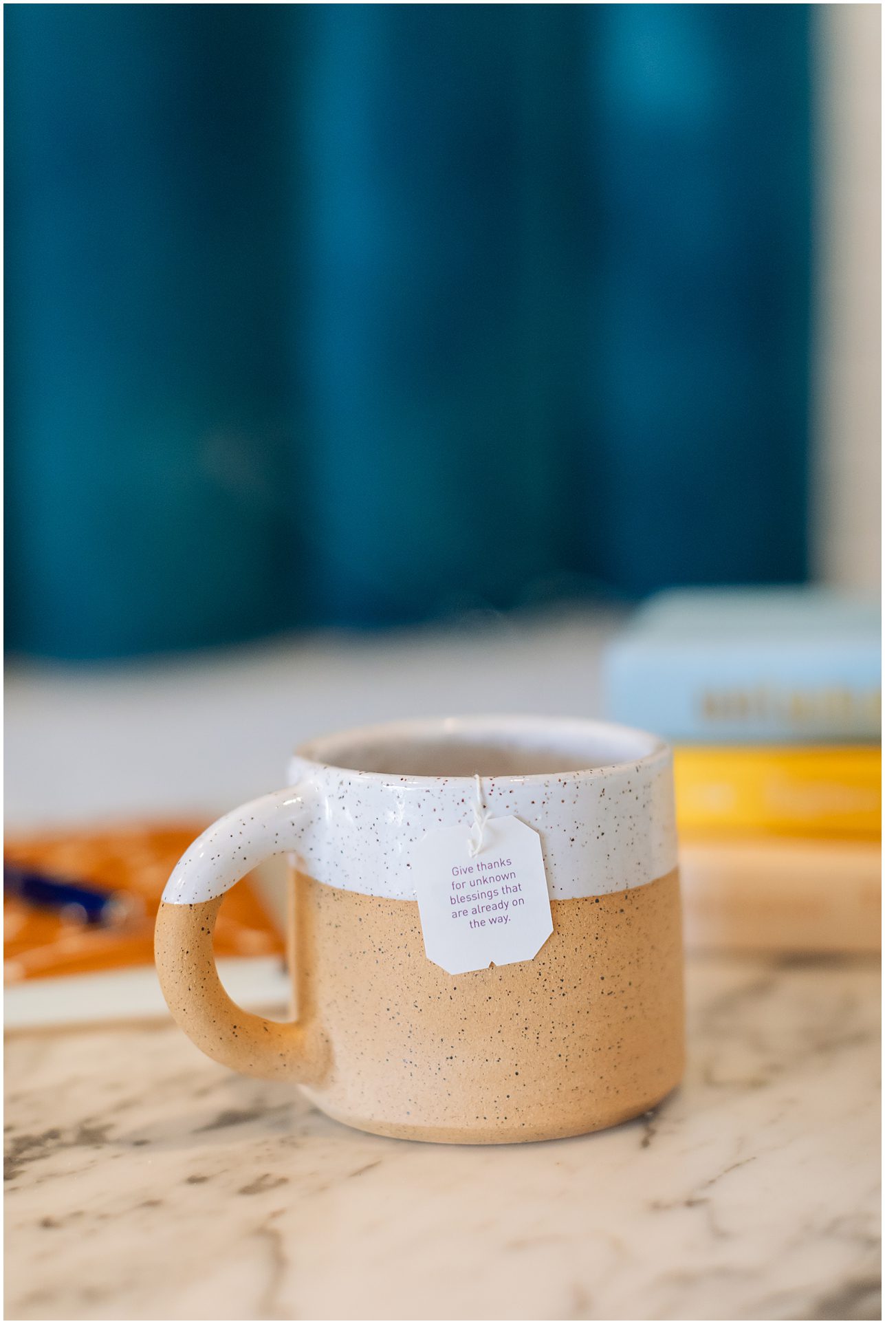A beige speckled mug with a tea bag on a marble surface, blue curtain and books in the background.