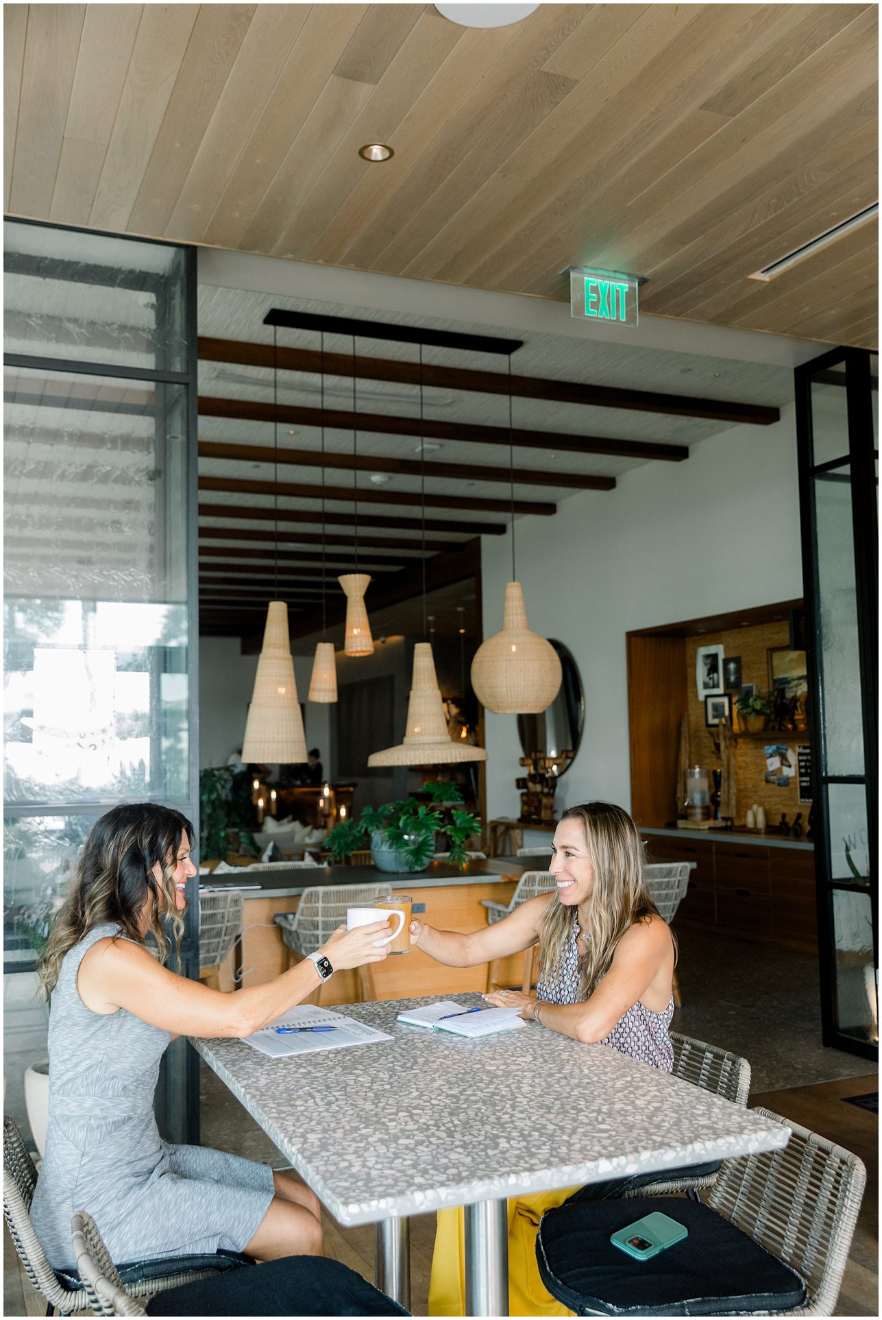 Two women sit at a table in a modern cafe, smiling and toasting coffee mugs together.