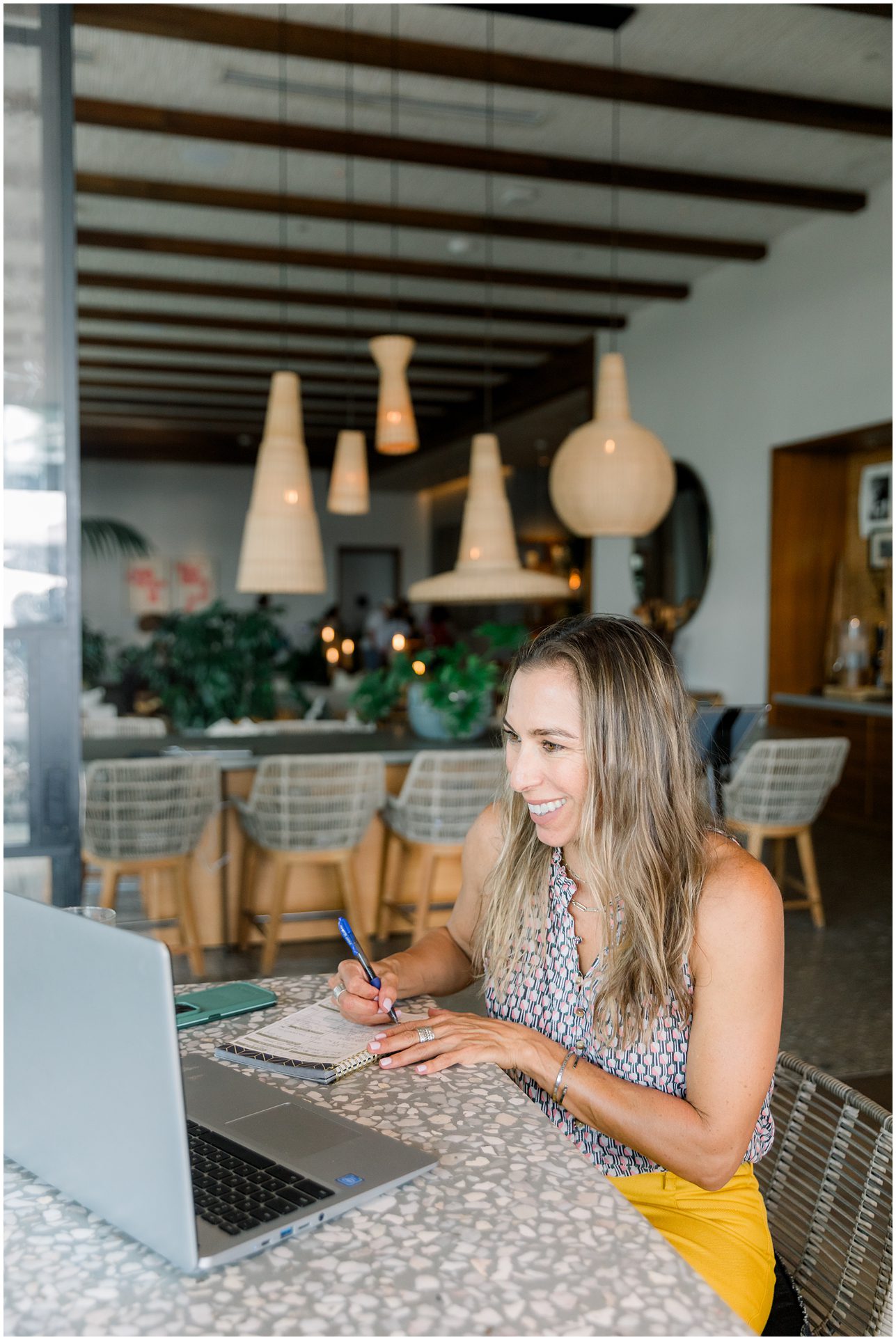 Woman smiling and working on a laptop at a modern, stylish cafe table with hanging lights in the background.