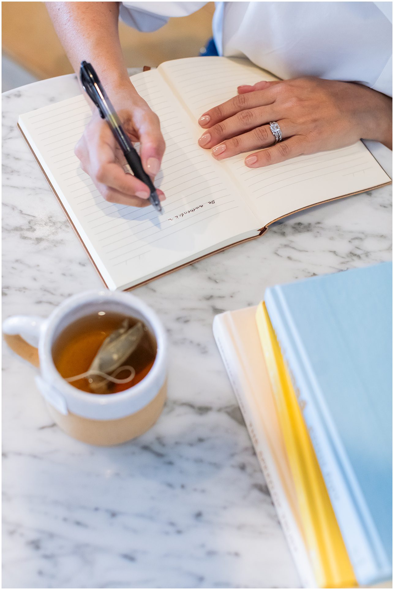 A person writes in a lined notebook on a marble table with tea and stacked books nearby.