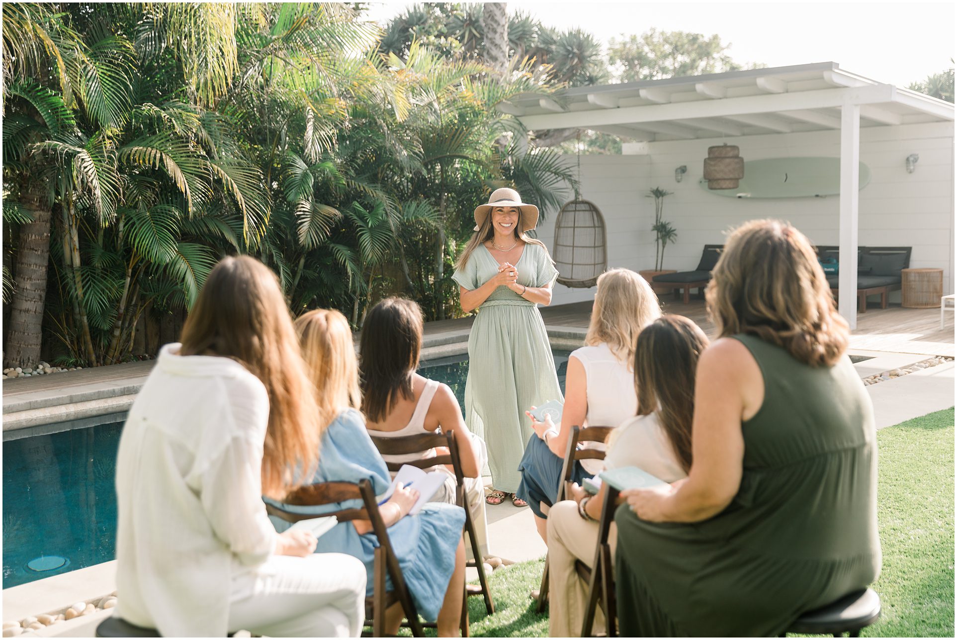 A woman in a hat speaks to a small group seated by a poolside, surrounded by greenery on a sunny day.