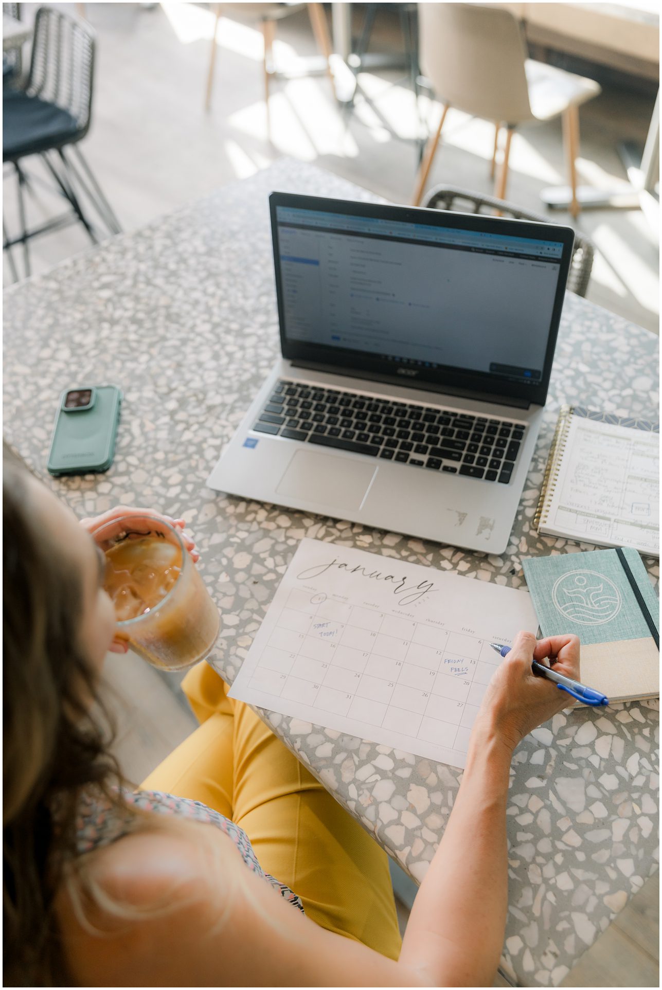 Person holding iced coffee, writing on a January calendar beside a laptop, notebook, and phone on a table.