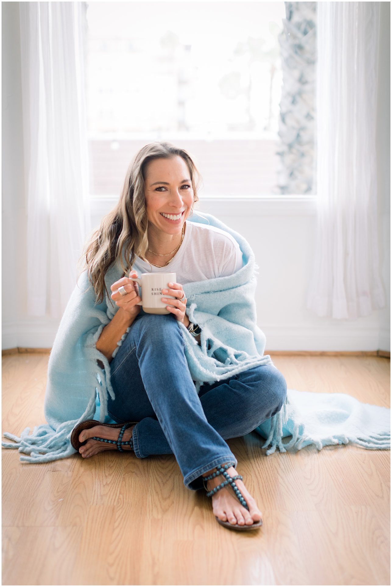 Woman sitting on the floor by a window, wrapped in a blue blanket, holding a mug and smiling.