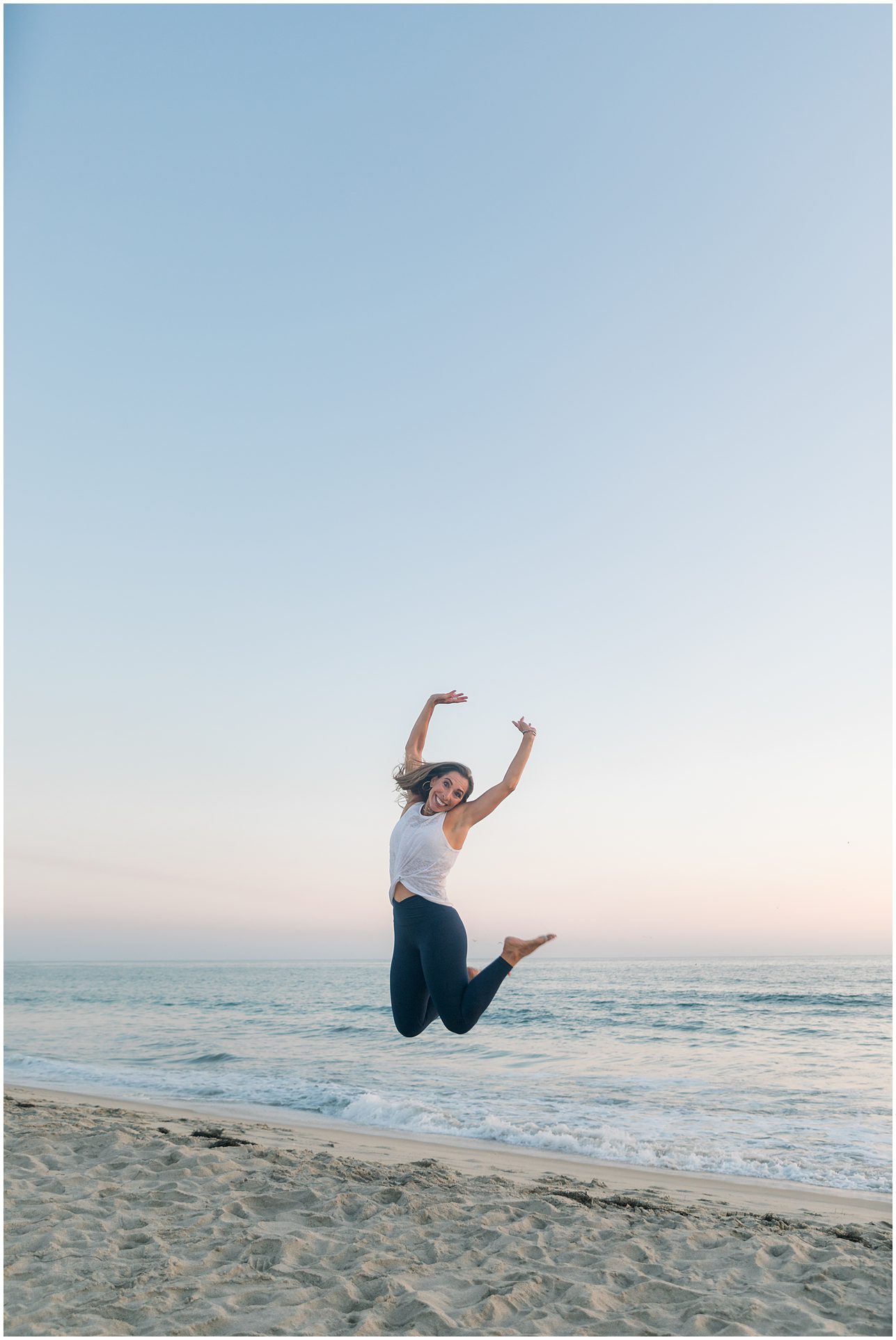 Woman jumping joyfully on a sandy beach at sunset with ocean waves and a clear sky in the background.