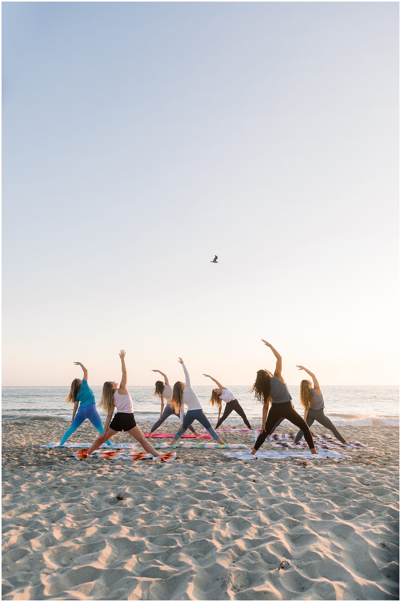 Seven people doing yoga on a sandy beach at sunrise or sunset, with the ocean and a bird in the background.